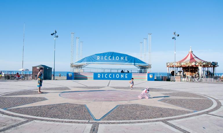 Piazza a Riccione con palco e giostra vicino al mare.