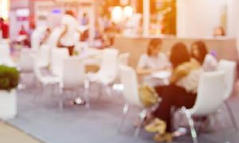 Fiera con persone sedute a tavoli bianchi, atmosfera luminosa.