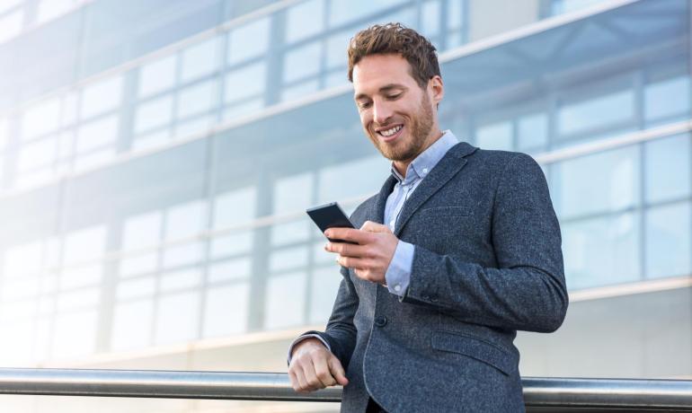 Uomo sorridente con smartphone davanti a edificio moderno.