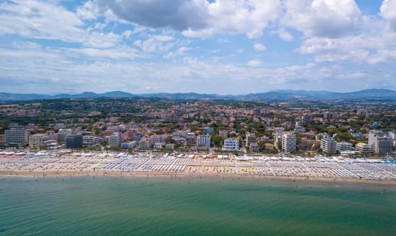 Spiaggia affollata con ombrelloni e mare limpido, città sullo sfondo.
