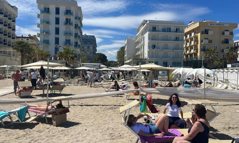 Spiaggia affollata con lettini e ombrelloni, edifici sullo sfondo.