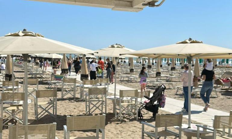 Spiaggia soleggiata, ombrelloni bianchi, bicchieri rossi, atmosfera vivace e gioiosa.