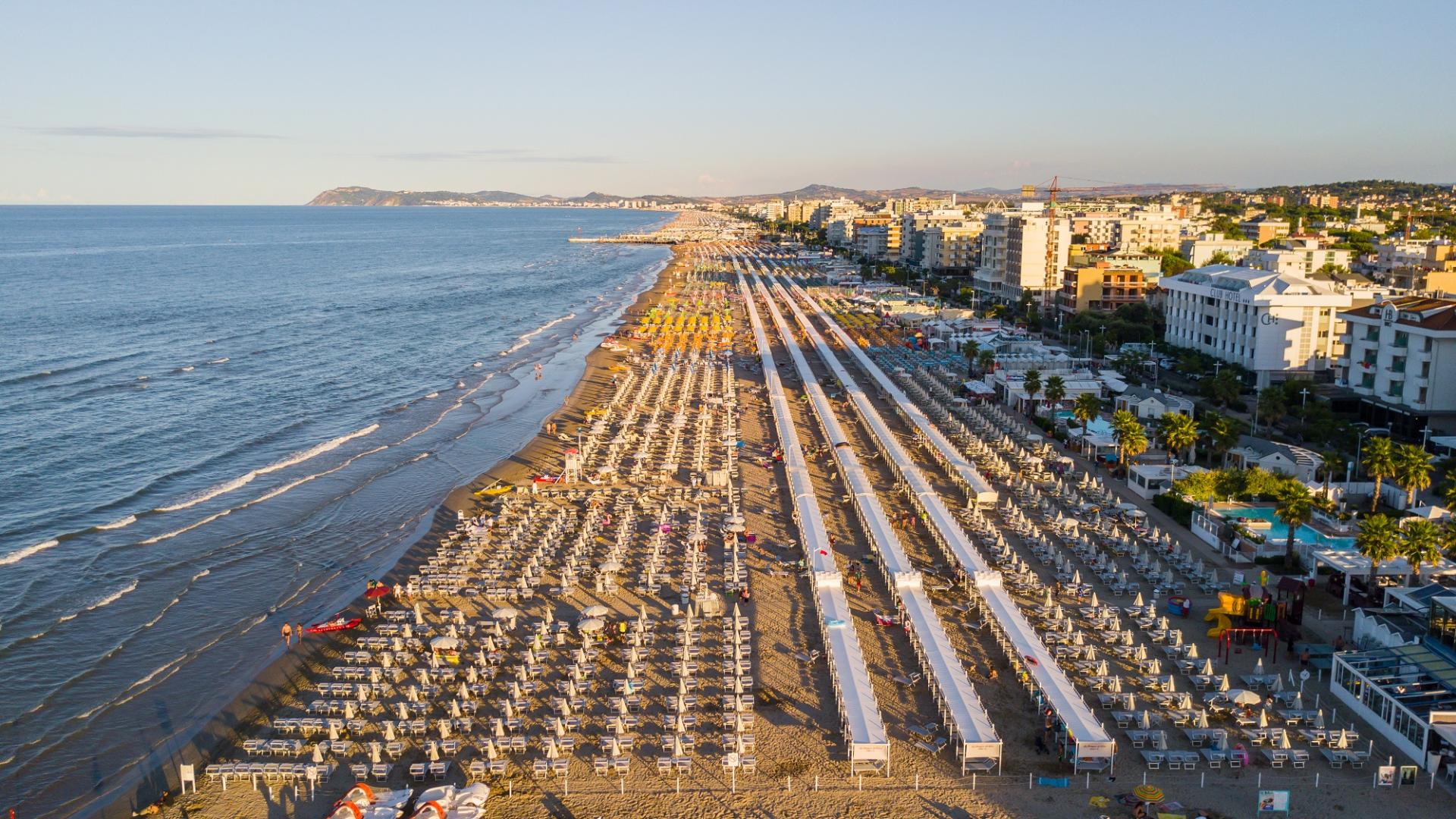 Spiaggia affollata con ombrelloni, piscina e pista ciclabile lungo la costa.