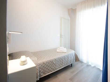 Camera minimalista con letto singolo, tende bianche e luce naturale.