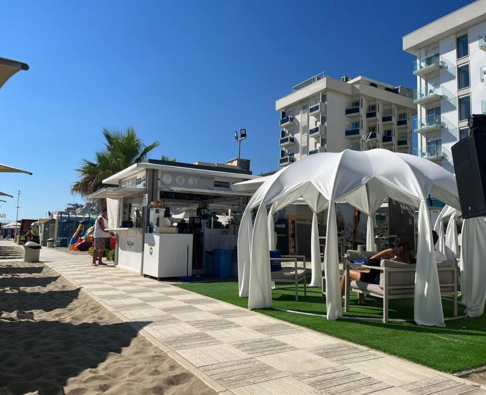 Spiaggia con bar, gazebo bianco e persone rilassate, edifici sullo sfondo.
